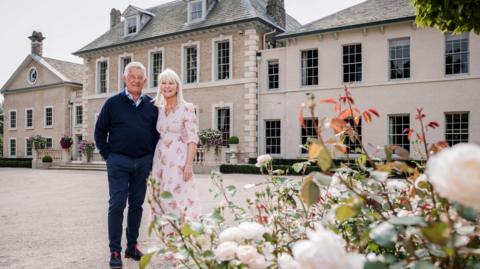 David and Linda Kilburn stand outside Hotham Hall in East Yorkshire. Mr Kilburn is wearing navy shoes, jeans and jumper with a light blue shirt. Mr Kilburn is wearing pink floral dress.