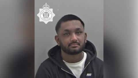 A police mugshot showing a man with very short dark hair with a dark beard wearing a white T-shirt and a black hoodie. 