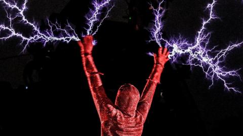 A person covered entirely by a red glittery body suit is pictured with their back to the camera with both arms raised in the air. Bright white-blue lighting bolts appear to burst from their hands against a background of total darkness.