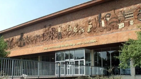 The front of a museum building. It has a large, brown brick facade with depictions of people and equipment on the front. There are wagons, people making pottery and large kilns on the front. In gold writing beneath it, a sign reads "The Potteries Museum and Art Gallery"