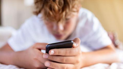 Fair-haired boy lying on his front looking at his mobile phone screen. 