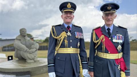 Two men in navy blue army suits. They are standing next to a stone statue of a person sitting down.