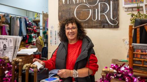 Julie Teckman is photographed standing at a shop till, smiling at the camera. She is wearing a red knitted jumper with a black, hooded bodywarmer. She is stood in front of a cream coloured wall with a big brown sign above her head saying "Guru". 