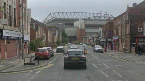A wide single carriage road leads to Anfield stadium. There are a number of parked cars in front of shops.