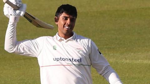Rishi Patel wearing cricket whites celebrates scoring a century by raising his bat