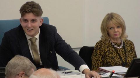 George Finch and Monica Fogarty sitting at a table during a meeting. Finch has short brown hair and is wearing a suit. He has his left hand on the table and is looking to his right while smiling. Fogarty is wearing a silver necklace and a yellow and black top. She is tilting her head to the right while looking towards Finch.