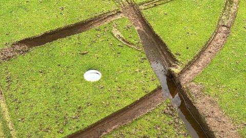 Picture of the green of one hole at the golf club. The white hole is pictured, surrounded by several deep grooves which are muddy and made by the tyres of motorbikes.
