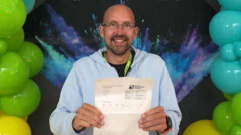 A bald man wearing a hoodie standing in front of a wall of multi-coloured balloons. He is holding up a piece of paper and smiling.