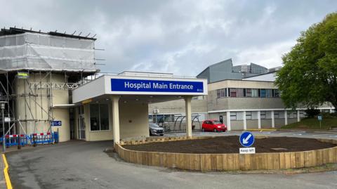 A view of Guernsey's Elizabeth Hospital showing the main entrance. It shows a single story building with scaffolding and cars. 