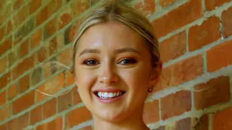 A young woman with blonde hair tied back. She is smiling at the camera and is stood in front of a brown brick wall
