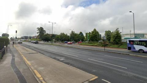 A street view image of the road shows several cars on either side. Trees line it in the background.