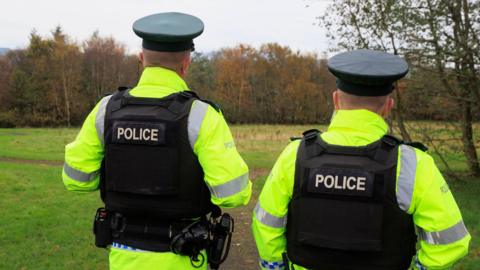 Two PSNI officers have their backs to the camera. They have on yellow police coats and black body armour that says 'police' on the back. 