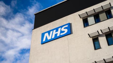 A grey building with an NHS logo on the side. We can see some windows. There is a blue sky behind it with some white clouds. 