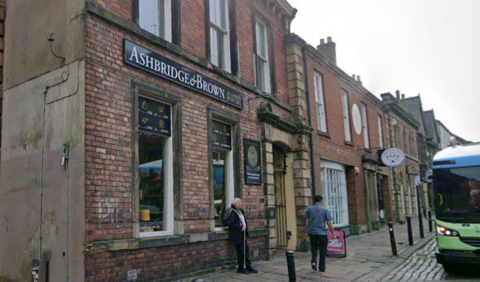 A Google Streetview screengrab showing the outside of Ashbridge & Brown in Carlisle city centre. The shop is in a old red-brick building with two tall and narrow windows on the front, where items are on display. There is a black shop sign with white and gold lettering and three people are walking past. The shop gives onto a cobbled street.