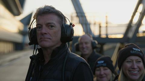 Graeme Lowdon stands in the pit at a race track. He is wearing large black headphones and a black jacket. A group of people stand behind him smiling. Two women are wearing black beanie hats with the Cadillac logo on them and a man who is slightly blurred and in the background is also wearing headphones