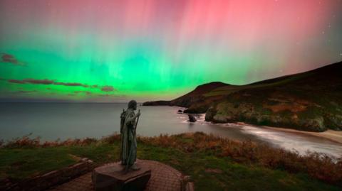 St. Crannog's statue is watching over the shoreline underneath the Northern Lights which is showcasing different colours such as green, pink and blue in the sky.