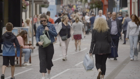 A general view of a busy hight street with people walking, some holding shopping bags. Many of the people in the background are out of focus. One woman, wearing cropped flowy black and white polka dot trousers and a denim jacket is wearing sunglasses and has a sage green rucksack held in front of her