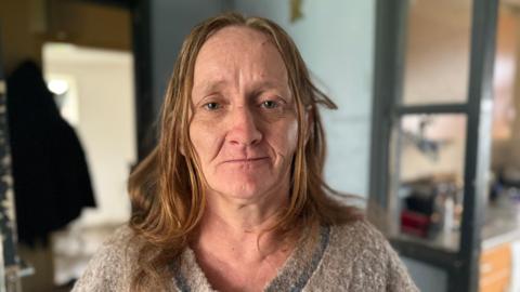 A woman with long auburn hair looks at the camera while standing inside a home, with the background out of focus. She is not smiling 