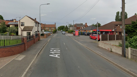 A Google streetview image of the A161 at Belton in North Lincolnshire. The road has a tarmac path on either side which is also lined with gardens and houses. A small convenience store, with red and blue branding, is visible on the right of the image.