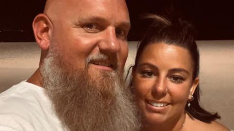 A man with a bald head and a long beard is next to a woman with dark brown hair. They are standing close to one another while posing for the photograph.