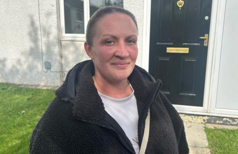 A woman stands in a garden in front of a black front door, weearing a bobbly dark fleece and a white t-shirt. Her hair is tied back and she is smiling while looking directly at the camera.