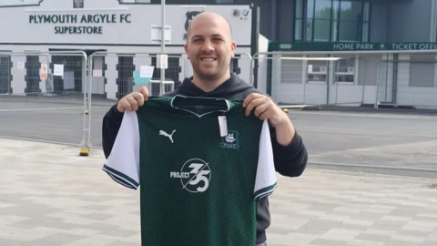 A person holds up a Plymouth Argyle shirt outside Home Park stadium, with the Plymouth Argyle FC Superstore visible in the background.