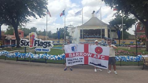 Three men hold up a large England flag with the words Barrow AFC across the horizontal line of the red cross. In the top left corner it has the Barrow AFC badge, the top right has a red rose, the bottom left has the phrase "you fill up my senses" and the remaining quarter has the outline of a black submarine. The three men are all wearing sunglasses and are standing in front of a sign for the theme park Six Flags Over Texas. There is a pavilion and a rollercoaster in the background.