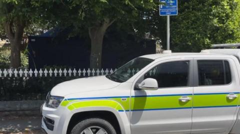 A white police vehicle with a yellow stripe outlined in blue. The vehicle is parked and has dark windows. There is a black and white fence behind with trees behind.