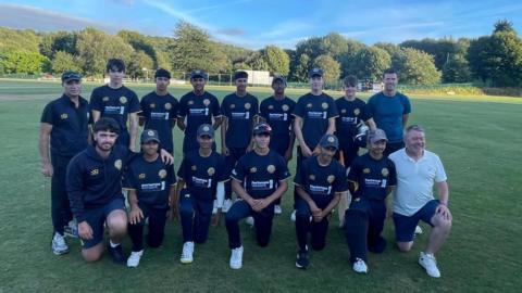 An 15 cricket team in navy sports gear and caps, posing with coaches