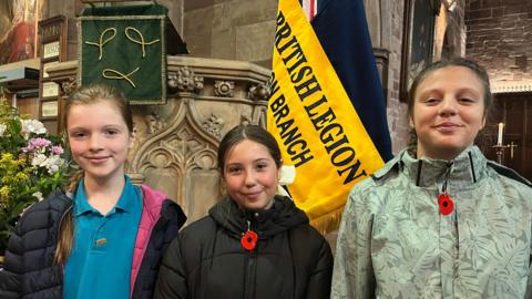 Three school girls are standing in front of a blue and yellow royal British legion flag in a church. Ella has brown hair tied in a plait. Maisie has brown hair and is wearing a red poppy. Lena has brown hair in two plaits and is wearing a red poppy.