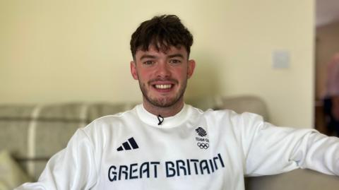 A smiling Ed Lowe seated on a sofa, wearing a white sweatshirt with "Great Britain" written across it