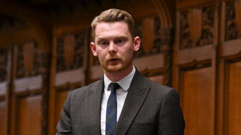 MP Luke Myer speaking in Parliament. He has short ginger hair combed to one side and a trimmed beard. He is wearing a grey suit with a white shirt and blue and green chequered tie. Behind him is brown wooden panelling with engraved decorations.