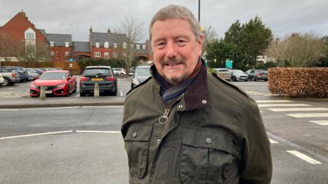 A man with grey hair, wearing a dark green jacket, smiles at the camera with hsi arms behind his back. A car park is behind him.