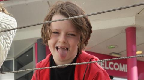 Lennix smiling at camera with his tongue out. He has long fair hair and is wearing a red top over a black T-shirt