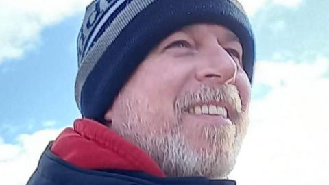 Corporal David Thorne, man smiling, looking away from camera, with a grey beard and wearing a blue and grey wool hat.