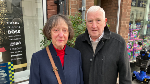 An elderly couple standing in front of a shop. The woman has brown hair, a red jumper and a blue coat and the man has white hair, a white shirt and a black coat.