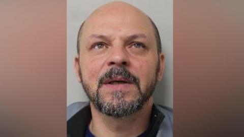 Mugshot of Nederi. He is bald with a round head and short black and grey beard