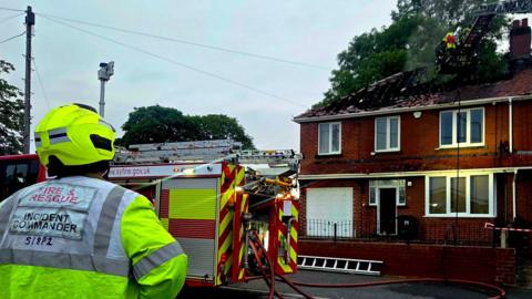 A fire engine outside a residential property, which has a burnt roof. A fire fighter wearing fluroscent clothing looks on.