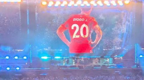 Giant screen tribute to Diogo Jota, He has his back to the camera and you can see a large 20 and his name Diogo J. on the back of his red shirt. In front of the large screen Oasis are playing a song.