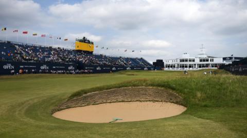 a golf course, with a sandy bunker in the foreground. On the left there is a large stand, approximately half full of fans. Above that, there is a small TV gantry for cameras. To the right of the stand, a large white building is pictured. On the course, there are several golf players and caddies in the distance.