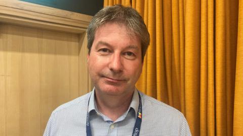 A man wearing a black and white shirt and a lanyard, is standing in front of an orange curtain.