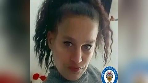 A woman with curly dark hair and black eyeliner, wearing a grey turtleneck, looks at the camera. The West Midlands Police logo is at the bottom right of the picture.