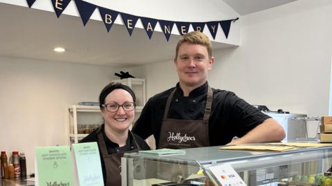 A man and a woman smile from behind the counter at the Beanery Cafe in Devizes, they wear black T-shirts and brown aprons with the company's logo.