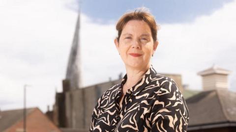 A woman with brown hair stood in front of the camera with Chesterfield's crooked spire in the background. 