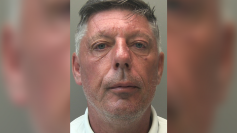 A police custody shot of Matthew Dunn looking at the camera. He is wearing a white polo shirt and has short grey hair. The wall behind him in grey.