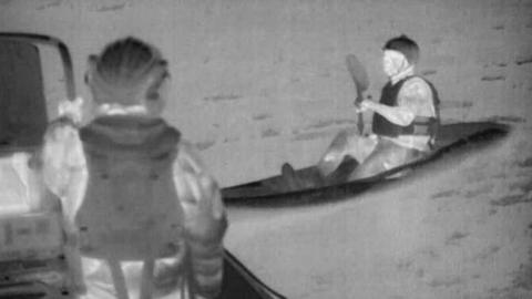 A black and white picture of two people, one kayaking and one standing on a boat. They are both in the water.