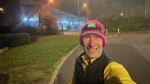 Photo shows Ted Winterton taking a selfie, he is wearing a bright yellow fleece underneath an armless black coat. He is wearing a bright pink hat with a headtorch on. He is smiling whilst running on a footpath by the side of an industrial park. 