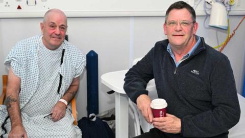 Two men sit side by side in a hospital. One is in a white plastic hospital gown after treatment.