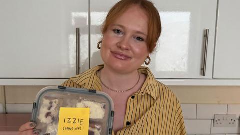 A woman holding a lunch box that has a sticker on it saying "Izzie (Hands off)"