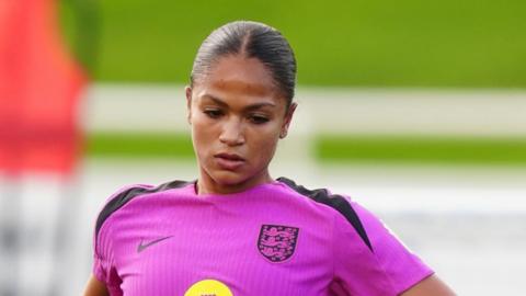 Taylor Hinds in a mauve England football training kit. Her hair is pulled back in a tight pony tail and she is looking downwards.